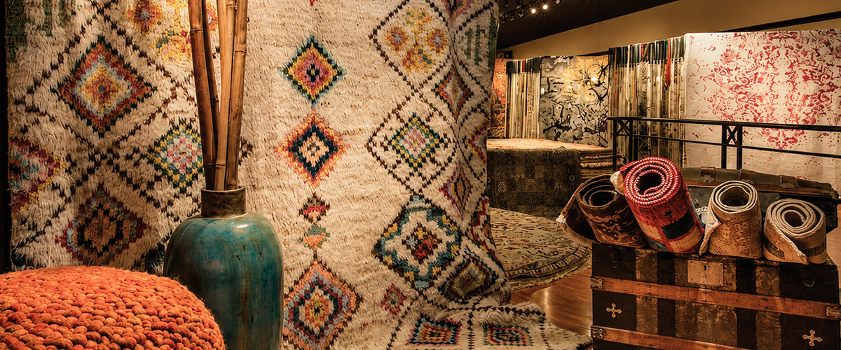 Showroom corner with Moroccan diamond rugs, rolled rugs, a teal vase and an orange pouf